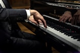 A person dressed in a black suit is playing a grand piano, focusing on the hands and keys. The sleeve has three buttons. The piano is a Steinway & Sons model with a shiny black surface reflecting the player’s hands.