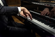 A person dressed in a black suit is playing a grand piano, focusing on the hands and keys. The sleeve has three buttons. The piano is a Steinway & Sons model with a shiny black surface reflecting the player’s hands.