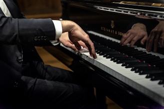 A person dressed in a black suit is playing a grand piano, focusing on the hands and keys. The sleeve has three buttons. The piano is a Steinway & Sons model with a shiny black surface reflecting the player’s hands.