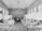 A long hospital ward with numerous beds lined up on both sides, each covered with white sheets. A nurse or medical professional in a hat and white uniform stands near the beds. The walls and ceiling are wooden, with a row of windows allowing natural light to enter the room. Vintage lighting fixtures hang from the ceiling, and there is a fireplace at the end of the room.