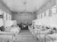 A long hospital ward with numerous beds lined up on both sides, each covered with white sheets. A nurse or medical professional in a hat and white uniform stands near the beds. The walls and ceiling are wooden, with a row of windows allowing natural light to enter the room. Vintage lighting fixtures hang from the ceiling, and there is a fireplace at the end of the room.
