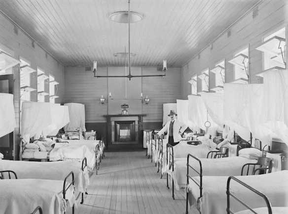 A long hospital ward with numerous beds lined up on both sides, each covered with white sheets. A nurse or medical professional in a hat and white uniform stands near the beds. The walls and ceiling are wooden, with a row of windows allowing natural light to enter the room. Vintage lighting fixtures hang from the ceiling, and there is a fireplace at the end of the room.