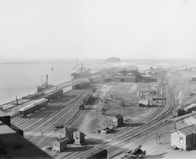 Black and white image of a cargo ship docked at a busy port with containers being loaded.