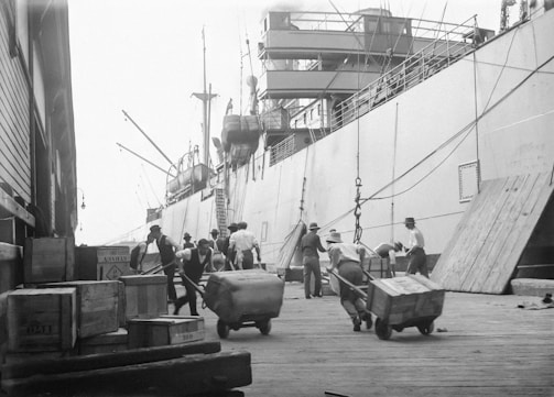 A bustling dock scene with several workers wearing hats and vests, moving large crates and cargo using handcarts. A large ship is docked alongside, with ropes and gangways connecting it to the pier. The background features the ship's deck and masts, while the foreground shows various crates stacked on the dock.