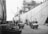 A bustling dock scene with several workers wearing hats and vests, moving large crates and cargo using handcarts. A large ship is docked alongside, with ropes and gangways connecting it to the pier. The background features the ship's deck and masts, while the foreground shows various crates stacked on the dock.