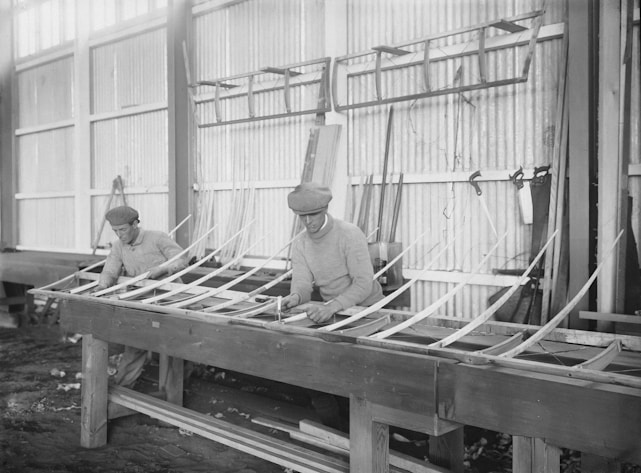 Two men in flat caps are constructing a wooden framework in a workshop. They are using traditional woodworking techniques, surrounded by tools and materials. The setting gives a historical feel with large windows and corrugated walls.