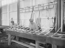 Two men in flat caps are constructing a wooden framework in a workshop. They are using traditional woodworking techniques, surrounded by tools and materials. The setting gives a historical feel with large windows and corrugated walls.