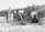 A group of women and a child are engaged in outdoor work, with some using shovels to dig in the sandy ground. The environment appears rural, with grassy vegetation and a clear sky. The women are dressed in long skirts and blouses, typical of an earlier time period.