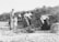A group of women and a child are engaged in outdoor work, with some using shovels to dig in the sandy ground. The environment appears rural, with grassy vegetation and a clear sky. The women are dressed in long skirts and blouses, typical of an earlier time period.