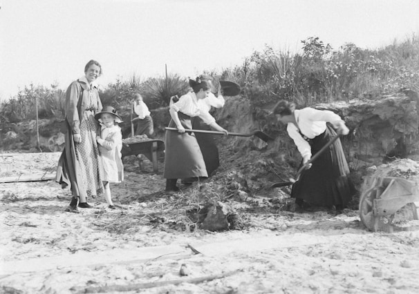 A group of women and a child are engaged in outdoor work, with some using shovels to dig in the sandy ground. The environment appears rural, with grassy vegetation and a clear sky. The women are dressed in long skirts and blouses, typical of an earlier time period.