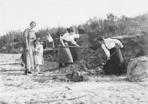 A group of women and a child are engaged in outdoor work, with some using shovels to dig in the sandy ground. The environment appears rural, with grassy vegetation and a clear sky. The women are dressed in long skirts and blouses, typical of an earlier time period.