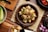 A wooden table features a bowl filled with a dish made of cubed tofu or similar food, garnished with chopped green herbs and sliced red onions. There are small bowls and plates nearby, some containing sliced red chili peppers and shallots. A cucumber is also visible on the side.
