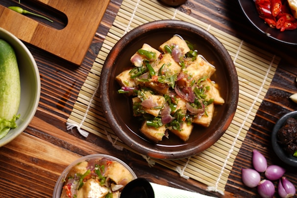 A wooden table features a bowl filled with a dish made of cubed tofu or similar food, garnished with chopped green herbs and sliced red onions. There are small bowls and plates nearby, some containing sliced red chili peppers and shallots. A cucumber is also visible on the side.