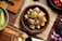 A wooden table features a bowl filled with a dish made of cubed tofu or similar food, garnished with chopped green herbs and sliced red onions. There are small bowls and plates nearby, some containing sliced red chili peppers and shallots. A cucumber is also visible on the side.