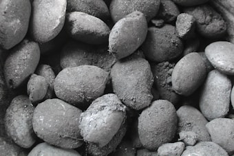 A close-up of numerous charcoal briquettes, with rough textures and varying shapes. The surface appears dusty and uneven, showcasing different sizes and orientations.