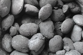 A close-up of numerous charcoal briquettes, with rough textures and varying shapes. The surface appears dusty and uneven, showcasing different sizes and orientations.