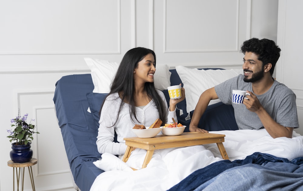 a man and a woman sitting on a bed with a tray of food