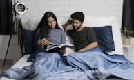 A couple is sitting on a bed, covered with blue blankets. The woman is showing something in a book to the man, and both seem engaged and smiling. There is a vintage-style floor lamp on the left and a small plant on a side table to the right. The background features a white brick wall.