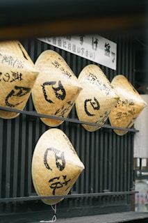 A row of traditional straw hats with black Japanese characters hang on a wooden fence-like structure. The texture of the woven material is visible, and a sign with more Japanese characters is placed above the hats.