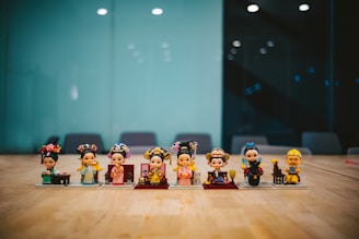 A row of eight small figurines is displayed on a wooden table, each dressed in traditional and colorful attire with detailed headdresses and accessories. They seem to be depicting characters from an Asian cultural theme. The background is a blurred office setting with a glass wall and dark chairs.