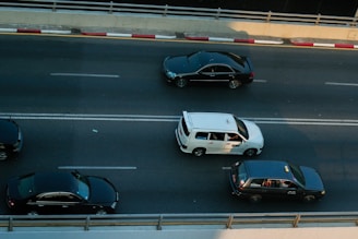 Three cars are traveling on a multi-lane road, with one white car in the center lane and two black cars in the adjacent lanes. The road is divided by double white lines and bordered by a concrete barrier with red and white markings.