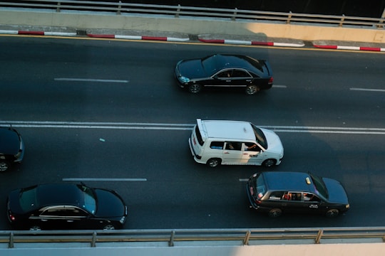Three cars are traveling on a multi-lane road, with one white car in the center lane and two black cars in the adjacent lanes. The road is divided by double white lines and bordered by a concrete barrier with red and white markings.
