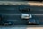 Three cars are traveling on a multi-lane road, with one white car in the center lane and two black cars in the adjacent lanes. The road is divided by double white lines and bordered by a concrete barrier with red and white markings.