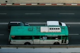 A green and white bus travels on a multi-lane road next to two cars, one black and the other white. The bus is marked with various writings on the side and appears slightly weathered. The road is bordered by a red and white striped curb.
