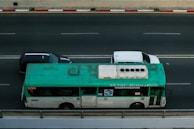 A green and white bus travels on a multi-lane road next to two cars, one black and the other white. The bus is marked with various writings on the side and appears slightly weathered. The road is bordered by a red and white striped curb.