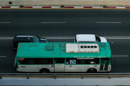 A green and white bus travels on a multi-lane road next to two cars, one black and the other white. The bus is marked with various writings on the side and appears slightly weathered. The road is bordered by a red and white striped curb.