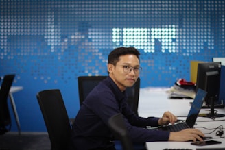 A person wearing glasses and a dark shirt sits at a desk, working on a laptop in a modern office environment. The background is a blue wall with a dot pattern that resembles a world map. Nearby, there are other desks with chairs and computers.