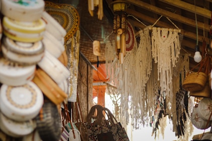A market stall displays various handmade crafts, including woven baskets, intricate macramé wall hangings, and embroidered hats. The setting is rustic, with wooden beams and ambient lighting, creating a cozy and artisanal atmosphere.