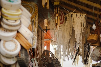 A market stall displays various handmade crafts, including woven baskets, intricate macramé wall hangings, and embroidered hats. The setting is rustic, with wooden beams and ambient lighting, creating a cozy and artisanal atmosphere.