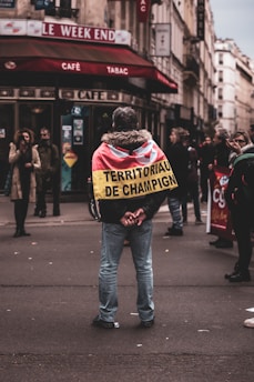 A person stands in the middle of a street facing away, wearing a jacket with a flag-like cloth draped over their shoulders. The text on the cloth reads 'TERRITORIAUX DE CHAMPIGNY'. Surrounding them are other pedestrians, some of whom appear to be gathered or engaged in conversation. The setting appears to be an urban area with a café named 'Le Week End' in the background.