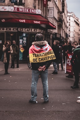 A person stands in the middle of a street facing away, wearing a jacket with a flag-like cloth draped over their shoulders. The text on the cloth reads 'TERRITORIAUX DE CHAMPIGNY'. Surrounding them are other pedestrians, some of whom appear to be gathered or engaged in conversation. The setting appears to be an urban area with a café named 'Le Week End' in the background.