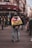 A person stands in the middle of a street facing away, wearing a jacket with a flag-like cloth draped over their shoulders. The text on the cloth reads 'TERRITORIAUX DE CHAMPIGNY'. Surrounding them are other pedestrians, some of whom appear to be gathered or engaged in conversation. The setting appears to be an urban area with a café named 'Le Week End' in the background.