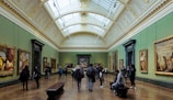 A spacious art gallery with ornate architectural elements and a high, arched ceiling filled with natural light. Several people are walking, standing, or sitting while observing large framed paintings on the green-painted walls.