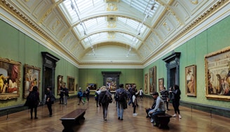 A spacious art gallery with ornate architectural elements and a high, arched ceiling filled with natural light. Several people are walking, standing, or sitting while observing large framed paintings on the green-painted walls.