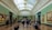A spacious art gallery with ornate architectural elements and a high, arched ceiling filled with natural light. Several people are walking, standing, or sitting while observing large framed paintings on the green-painted walls.