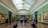 A spacious art gallery with ornate architectural elements and a high, arched ceiling filled with natural light. Several people are walking, standing, or sitting while observing large framed paintings on the green-painted walls.