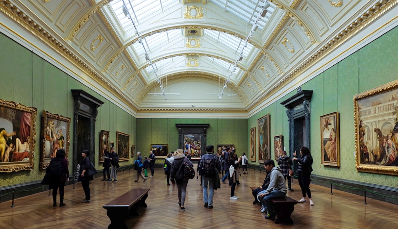 A spacious art gallery with ornate architectural elements and a high, arched ceiling filled with natural light. Several people are walking, standing, or sitting while observing large framed paintings on the green-painted walls.