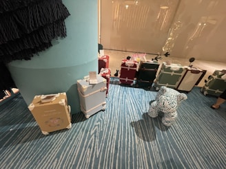 A collection of vintage-style suitcases is arranged on a blue, patterned carpet. The suitcases are in various colors, including beige, light blue, red, and green, and feature white accents and labels. A plush figure with patterns and text is positioned in front of the suitcases. The setting appears to be indoors, possibly a store or showroom, with a large column visible on the left side.