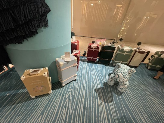 A collection of vintage-style suitcases is arranged on a blue, patterned carpet. The suitcases are in various colors, including beige, light blue, red, and green, and feature white accents and labels. A plush figure with patterns and text is positioned in front of the suitcases. The setting appears to be indoors, possibly a store or showroom, with a large column visible on the left side.