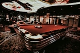 A luxurious interior features a billiard table with balls arranged on it. The ceiling has an intricate stained glass design, and the space is dimly lit, creating a sophisticated atmosphere. Comfortable chairs are placed around the area, with some decorative plants and a lamp adding to the elegance.