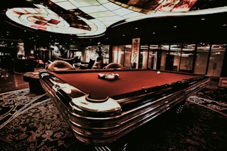 A luxurious interior features a billiard table with balls arranged on it. The ceiling has an intricate stained glass design, and the space is dimly lit, creating a sophisticated atmosphere. Comfortable chairs are placed around the area, with some decorative plants and a lamp adding to the elegance.