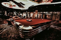 A luxurious interior features a billiard table with balls arranged on it. The ceiling has an intricate stained glass design, and the space is dimly lit, creating a sophisticated atmosphere. Comfortable chairs are placed around the area, with some decorative plants and a lamp adding to the elegance.