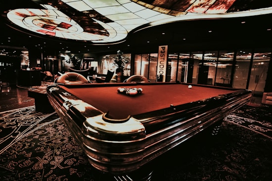 A luxurious interior features a billiard table with balls arranged on it. The ceiling has an intricate stained glass design, and the space is dimly lit, creating a sophisticated atmosphere. Comfortable chairs are placed around the area, with some decorative plants and a lamp adding to the elegance.