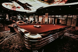 A luxurious interior features a billiard table with balls arranged on it. The ceiling has an intricate stained glass design, and the space is dimly lit, creating a sophisticated atmosphere. Comfortable chairs are placed around the area, with some decorative plants and a lamp adding to the elegance.