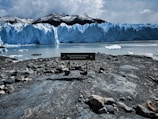 A large glacier with jagged icy peaks dominates the view. The glacier's blue hues contrast with the dark mountainous background. Foreground shows a rocky shoreline with a sign indicating the location as Parque Nacional Los Glaciares. Calm water reflects the glacier and the sky is overcast.