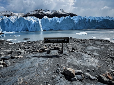A large glacier with jagged icy peaks dominates the view. The glacier's blue hues contrast with the dark mountainous background. Foreground shows a rocky shoreline with a sign indicating the location as Parque Nacional Los Glaciares. Calm water reflects the glacier and the sky is overcast.